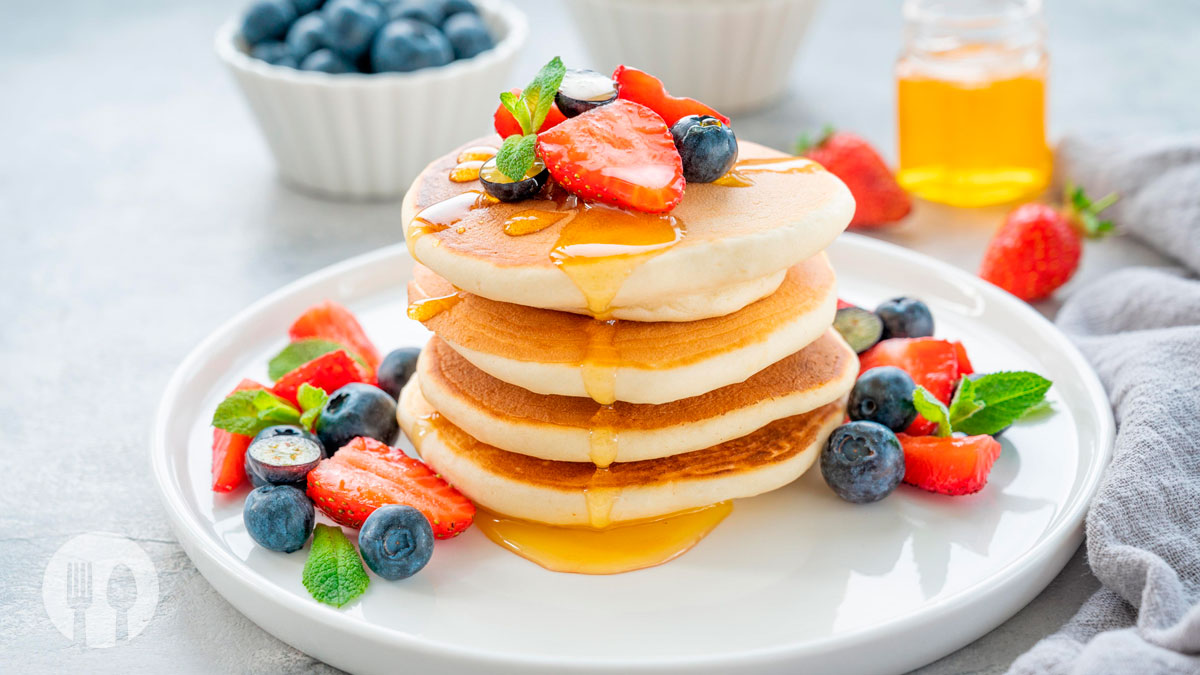 Panquecas caseiras fofinhas empilhadas num prato, acompanhadas de fruta fresca e mel.